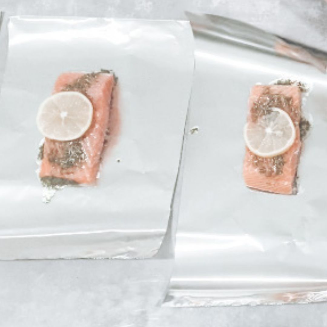 two pieces of salmon topped with a slice of lemon and herbs sitting on foil