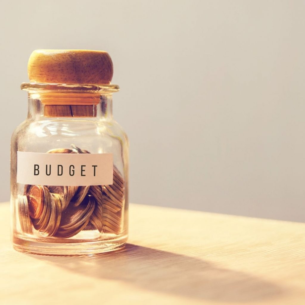 small glass jar with a cork lid that has coins inside and a white label with black words that say budget